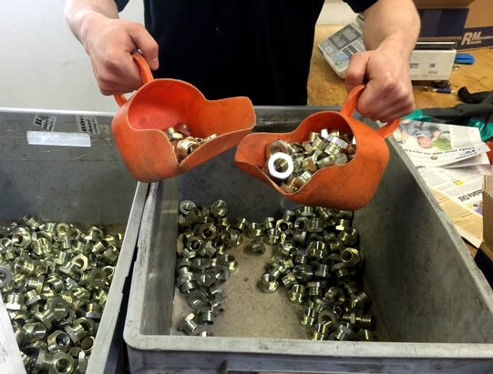 En person häller maskinbearbetade metalldelar (muttrar och kopplingar) från orange skopor ner i grå plastlådor på en arbetsyta i en verkstad.