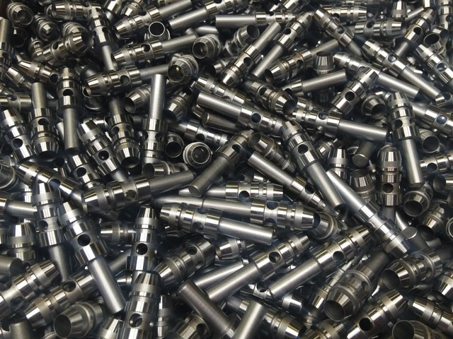 Närbild ovanifrån på ett stort antal CNC-svarvade metallkomponenter med cylindrisk form och räfflad yta, staplade löst på hög.