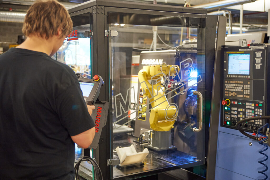 Operatör med ryggen mot kameran programmerar en gul Fanuc-robot med teach pendant framför en Doosan CNC-svarv i en MARAB-robotcell med glasdörr.