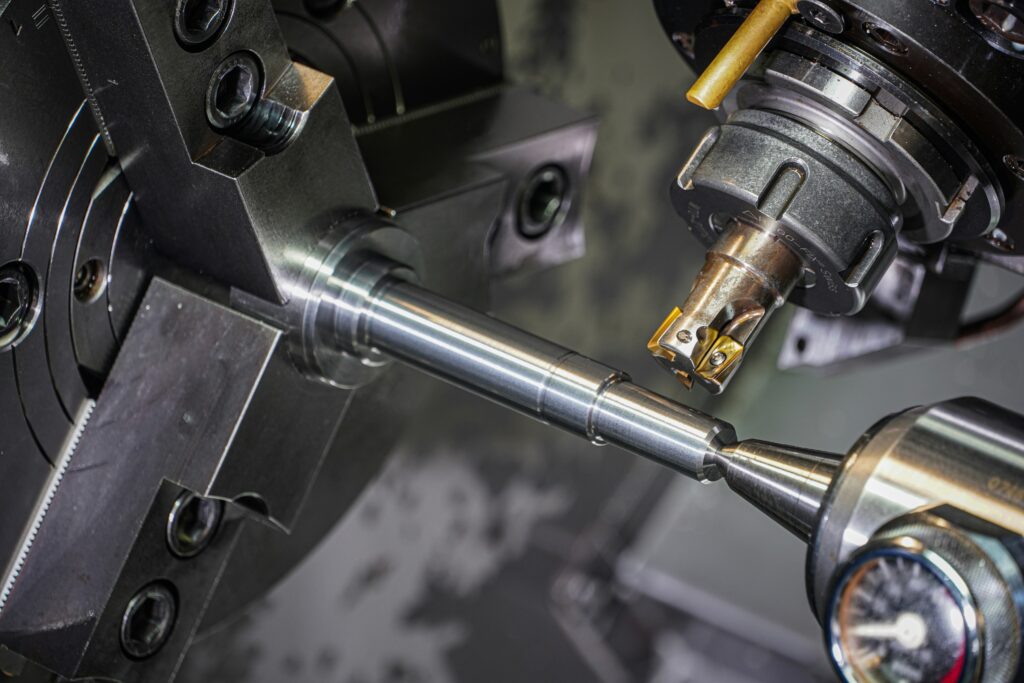 Närbild på CNC-svarvning där ett långt cylindriskt metallämne bearbetas samtidigt av två skärverktyg med guldfärgade hårdmetallskär, fastsatt i ett svart chuckfäste.